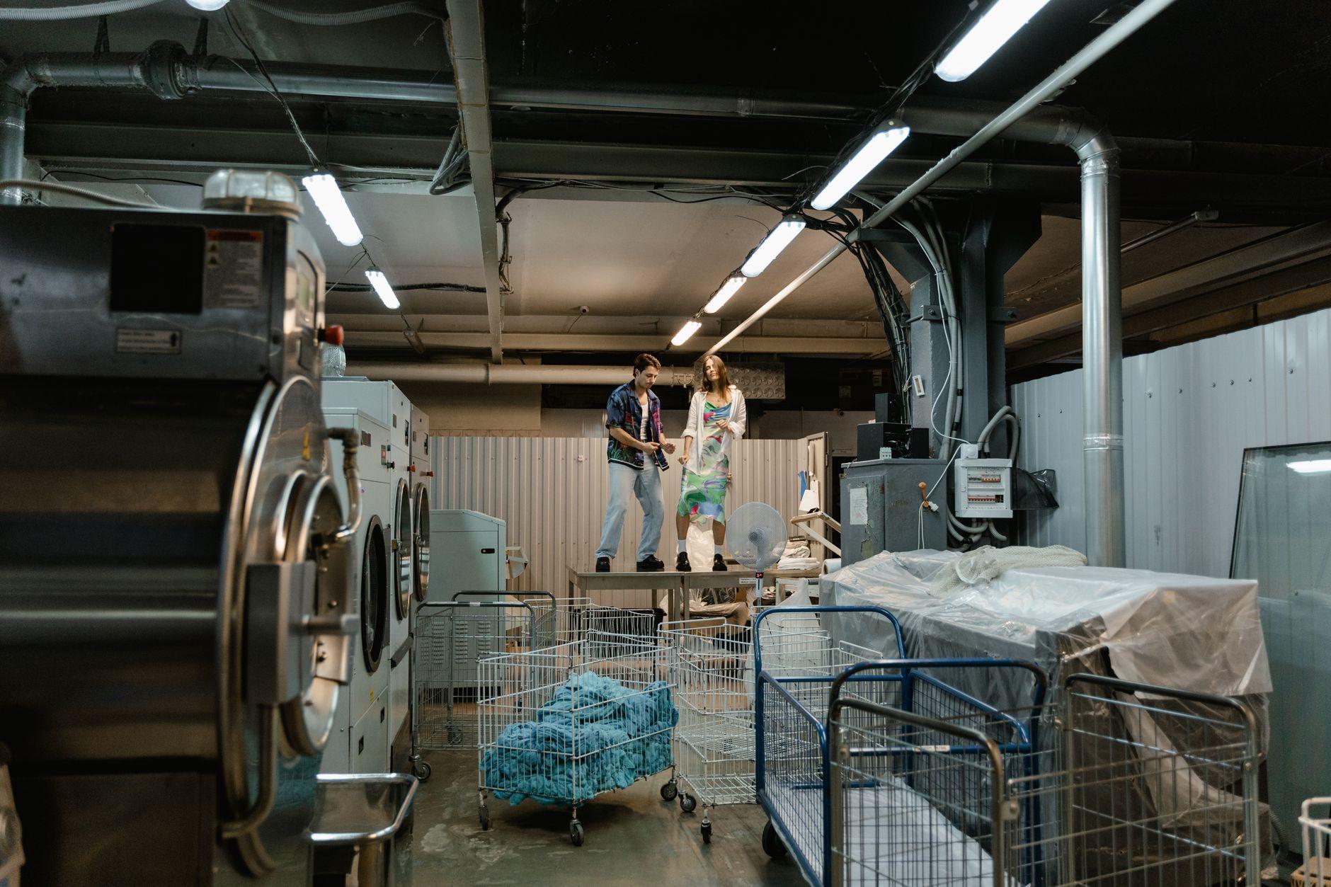 Two people working inside an industrial laundry facility with large machines and laundry carts.